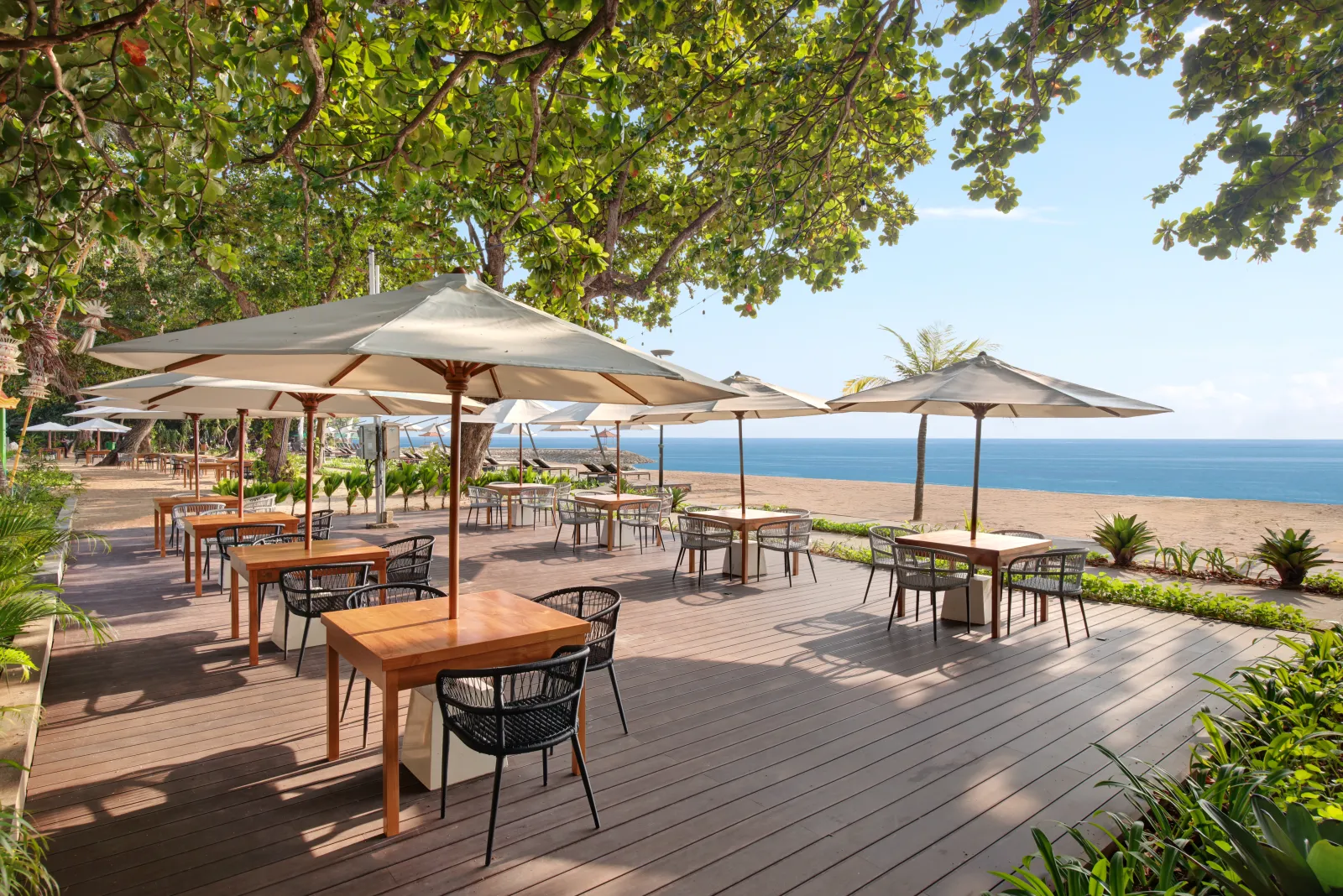 The beachside restaurant deck at éL Hotel & Resort Bali, teak tables under white umbrellas opening onto the sand and the sea at Sanur